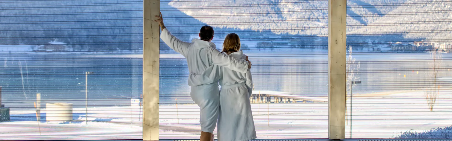 Zwei Personen in weißen Bademänteln blicken auf eine winterliche Landschaft.